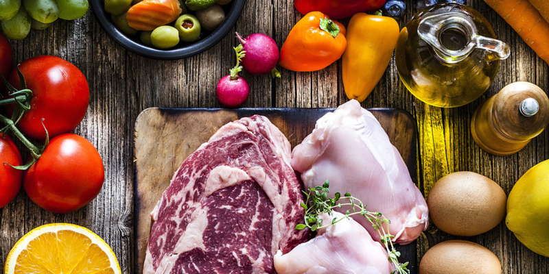 Food backgrounds: top view of a rustic wooden table filled with different types of food. At the center of the frame is a cutting board with beef steak, raw chicken meat and salmon fillet and all around it is a large variety of food like fruits, vegetables, cheese, bread, eggs, legumes, olive oil and nuts. DSRL studio photo taken with Canon EOS 5D Mk II and Canon EF 70-200mm f/2.8L IS II USM Telephoto Zoom Lens