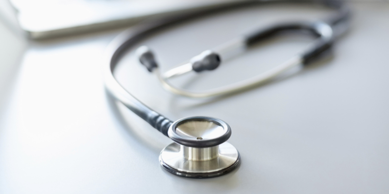 Close-up of stethoscope on desk with doctor's hands using laptop. The female doctor is sitting at desk in office. The focus is on stethoscope.