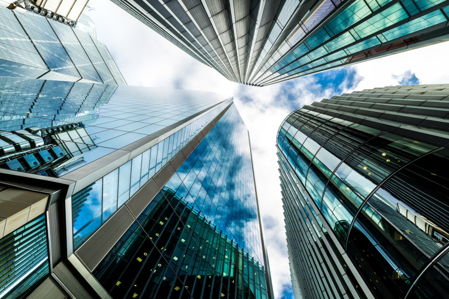 Wide angle image depicting an abstract view looking up at various different modern buildings and futuristic skyscrapers in central London. We can see a dazzling blue sky and cloudscape beyond. Room for copy space.