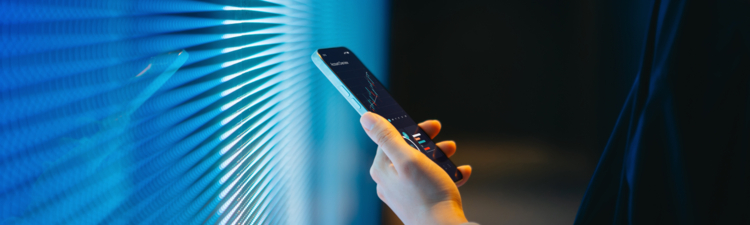 Cropped shot of young Asian woman making financial plans and analyzing investment trading data on smartphone against blue coloured illuminated LED digital display screen. Connecting to the future. Artificial intelligence. Finance and investment concept