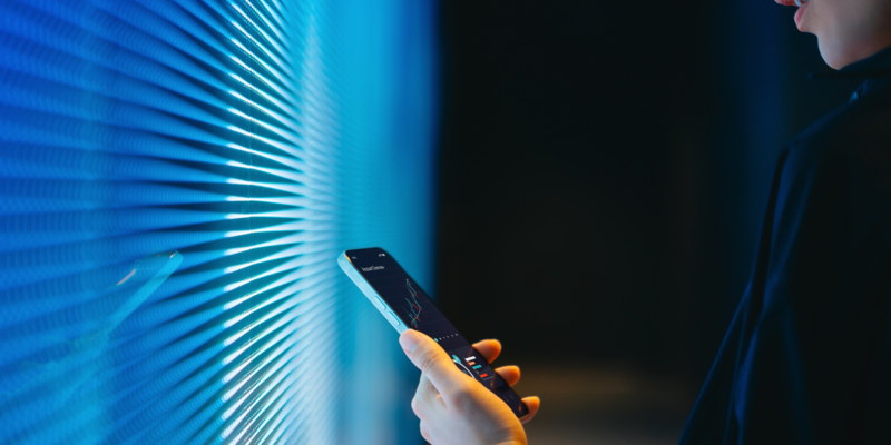 Cropped shot of young Asian woman making financial plans and analyzing investment trading data on smartphone against blue coloured illuminated LED digital display screen. Connecting to the future. Artificial intelligence. Finance and investment concept