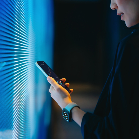 Cropped shot of young Asian woman making financial plans and analyzing investment trading data on smartphone against blue coloured illuminated LED digital display screen. Connecting to the future. Artificial intelligence. Finance and investment concept